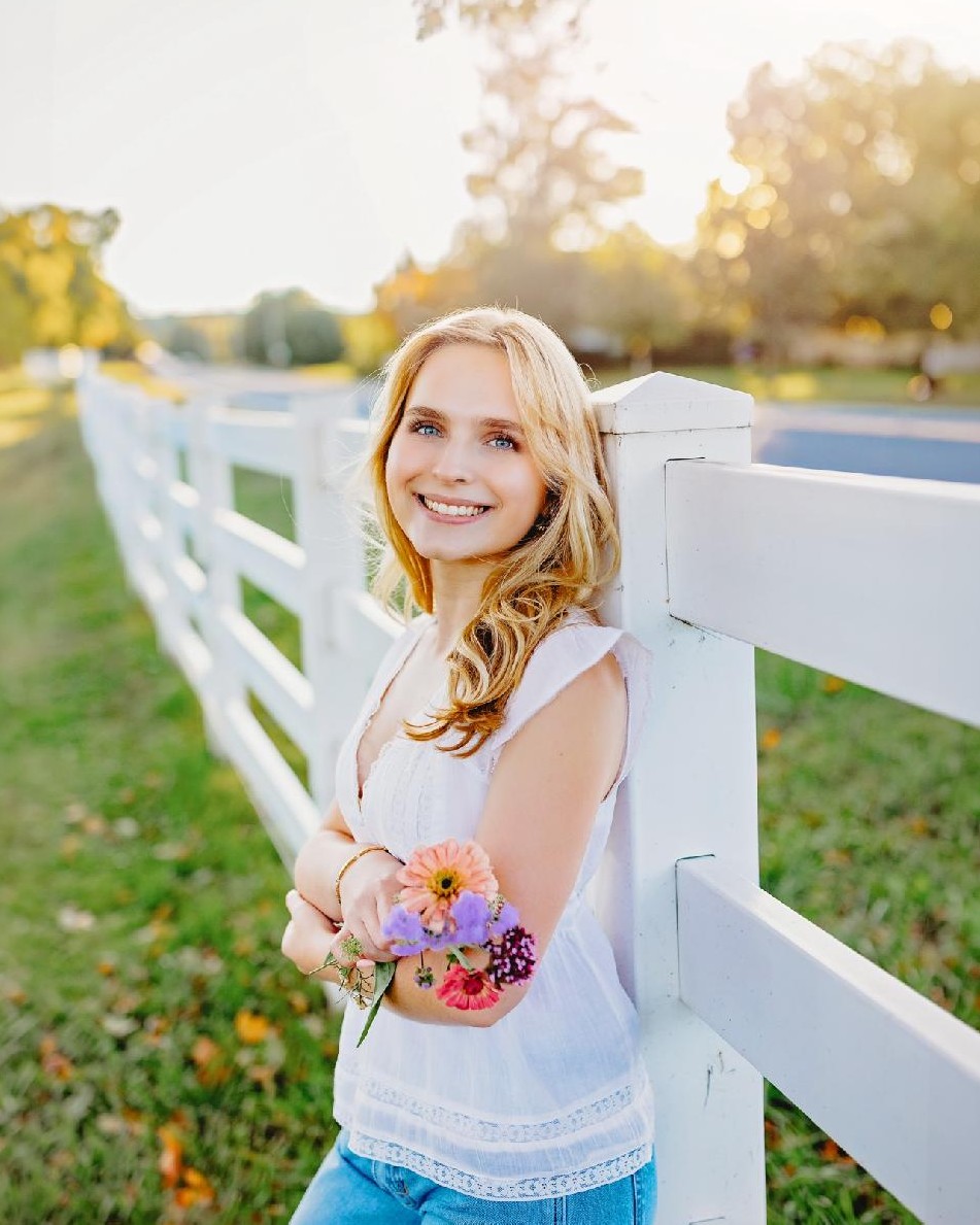 Madelyn Wright smiling at golden hour, leaning on a white fence and holding a small bouquet of zinnias